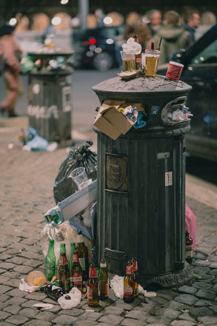 A black cylindrical public waste bin with a textured finish, situated on a cobbled street, is topped with various discarded items including plastic cups with foam lids and red straws, a crumpled paper bag, and a small cardboard box. Surrounding the base are multiple empty glass bottles and scattered litter, such as used napkins and scraps of paper. In the background, blurred figures of people and parked cars are visible, indicating a busy urban area. The scene suggests the bin has become overfilled, with trash spilling over onto the street surface. The environment appears to be a lively outdoor setting, possibly a marketplace or a busy town center, where rubbish removal by private services like Waste Removal Putney may be needed to manage refuse efficiently and maintain cleanliness.