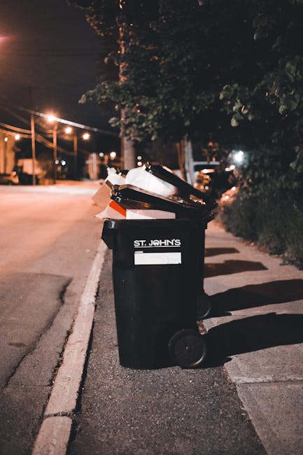A black wheeled rubbish bin with the label 'ST. JOHN'S' positioned on a concrete sidewalk beside a street at night. The lid of the bin is open, revealing scattered cardboard, paper, and plastic waste inside and protruding over the top of the bin. The surrounding environment includes a row of trees and bushes on the right, casting shadows on the pavement, with streetlights illuminating the scene with a warm glow. In the background, there are parked cars and distant streetlights, typical of an urban residential area. This scene suggests an instance of private waste disposal, possibly related to alternative rubbish collection services, with Waste Removal Putney capable of managing such waste clearance. The image reflects a typical scene for on-site rubbish disposal, where waste is temporarily stored outside a property in preparation for collection or removal, aligned with the principles of independent rubbish clearance.