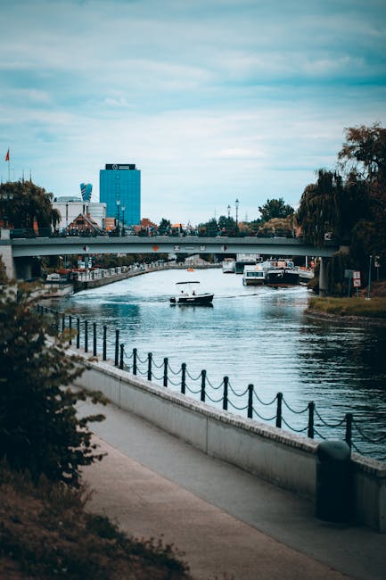 A view of a river running through an urban area, with a paved walkway and a decorative black chain fence running along the riverbank in the foreground. Several boats, including a small motorboat, are floating on the calm water. On the opposite side of the river, there are trees and green spaces, with a mix of modern and traditional buildings, including a prominent blue high-rise and a smaller white building with a red flag. A bridge crosses the river in the mid-distance, with additional boats docked along the waterfront. The sky is partly cloudy with a soft light illuminating the scene, indicating a clear day. The overall scene suggests an environment where private or independent waste disposal services, such as those provided by Waste Removal Putney, could be involved in managing rubbish removal from the surrounding urban and riverside areas, supporting alternative waste handling and on-site clearance activities.