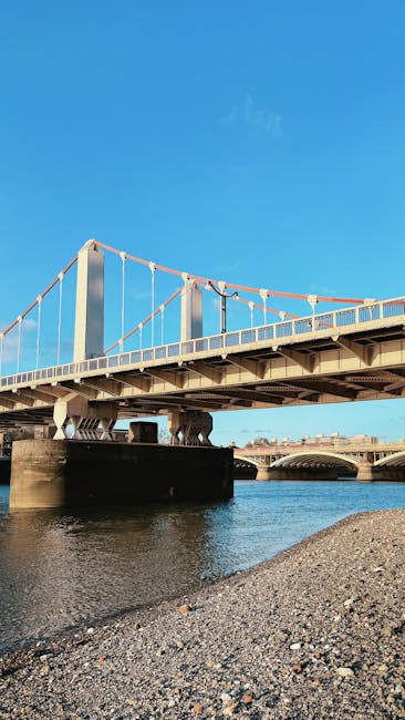 A large suspension bridge spans across a river, with tall vertical supports and cables connected in a swinging pattern. The bridge deck is visible, with metal railings along the edges and a smooth, light-colored surface. In the foreground, a gravelly riverbank with small stones and pebbles extends towards the water, which is calm with slight ripples. In the background, the opposite side of the river features a contemporary building with a curved roof, while the sky above is clear and bright blue, indicating a sunny day. The scene, as seen from below the bridge near the water’s edge, reflects a typical urban landscape that could relate to services such as private waste management or on-site clearance for areas around bridges and waterways, highlighting the environment where rubbish removal or clearance services may be required, as provided by Waste Removal Putney.