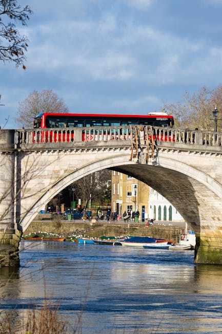A view of a river running through an urban area, with a paved walkway and a decorative black chain fence running along the riverbank in the foreground. Several boats, including a small motorboat, are floating on the calm water. On the opposite side of the river, there are trees and green spaces, with a mix of modern and traditional buildings, including a prominent blue high-rise and a smaller white building with a red flag. A bridge crosses the river in the mid-distance, with additional boats docked along the waterfront. The sky is partly cloudy with a soft light illuminating the scene, indicating a clear day. The overall scene suggests an environment where private or independent waste disposal services, such as those provided by Waste Removal Putney, could be involved in managing rubbish removal from the surrounding urban and riverside areas, supporting alternative waste handling and on-site clearance activities.