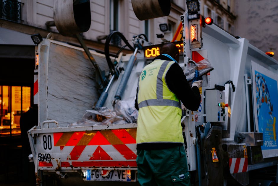 A sanitation worker wearing a high-visibility yellow vest and dark clothing is operating the controls of a large rubbish collection truck on a city street during dusk or early evening. The truck's rear is open, revealing a compartment filled with various waste materials, including crumpled paper and plastic. The vehicle has red and white reflective markings along its edges and illuminated warning lights on the back, indicating active loading or disposal. In the background, a building with warm interior lighting and large windows is visible, contributing to the urban setting. The image captures the moment of an independent collection service, such as Waste Removal Putney, conducting on-site waste clearance, illustrating the professional handling of refuse in a city environment at low lighting levels, with focus on the machinery, waste, and operational context.